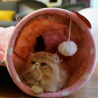 Pink plush cat tunnel bed with circular entrance, hanging pom-pom, and resting cat