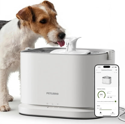 White pet water fountain with top nozzle; dog drinking while a phone shows controls.