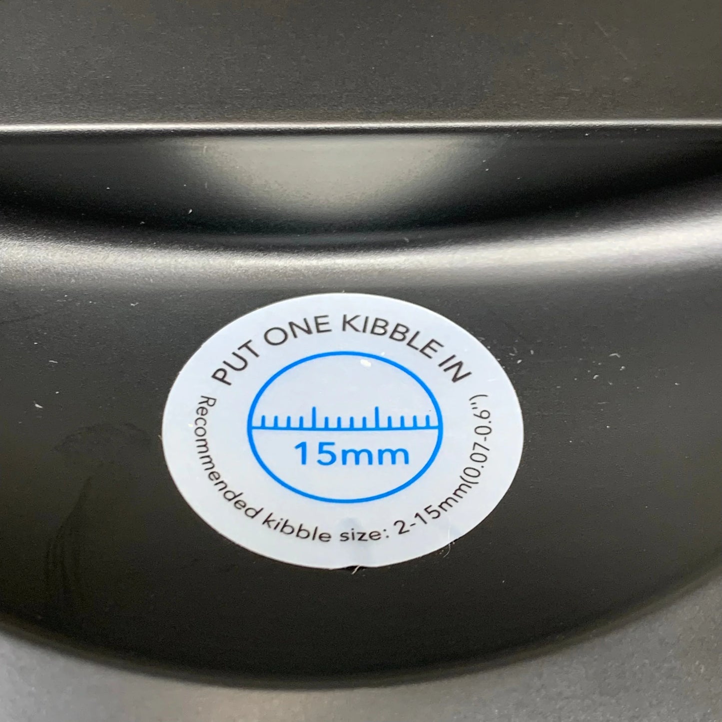 Black feeder lid label reading “PUT ONE KIBBLE IN” and 15mm guidance